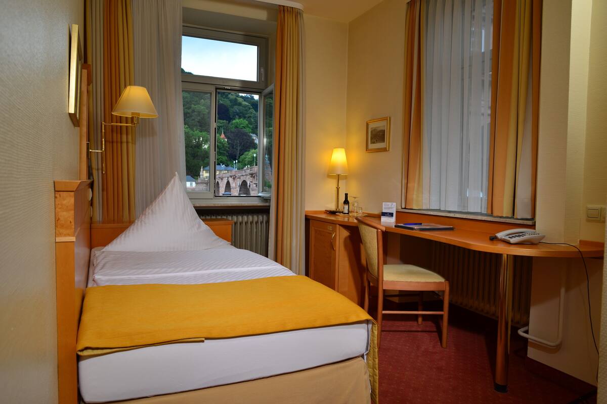 A hotel room with a bed, a desk with a chair, a lamp, a window with curtains, and a view of a bridge and trees.