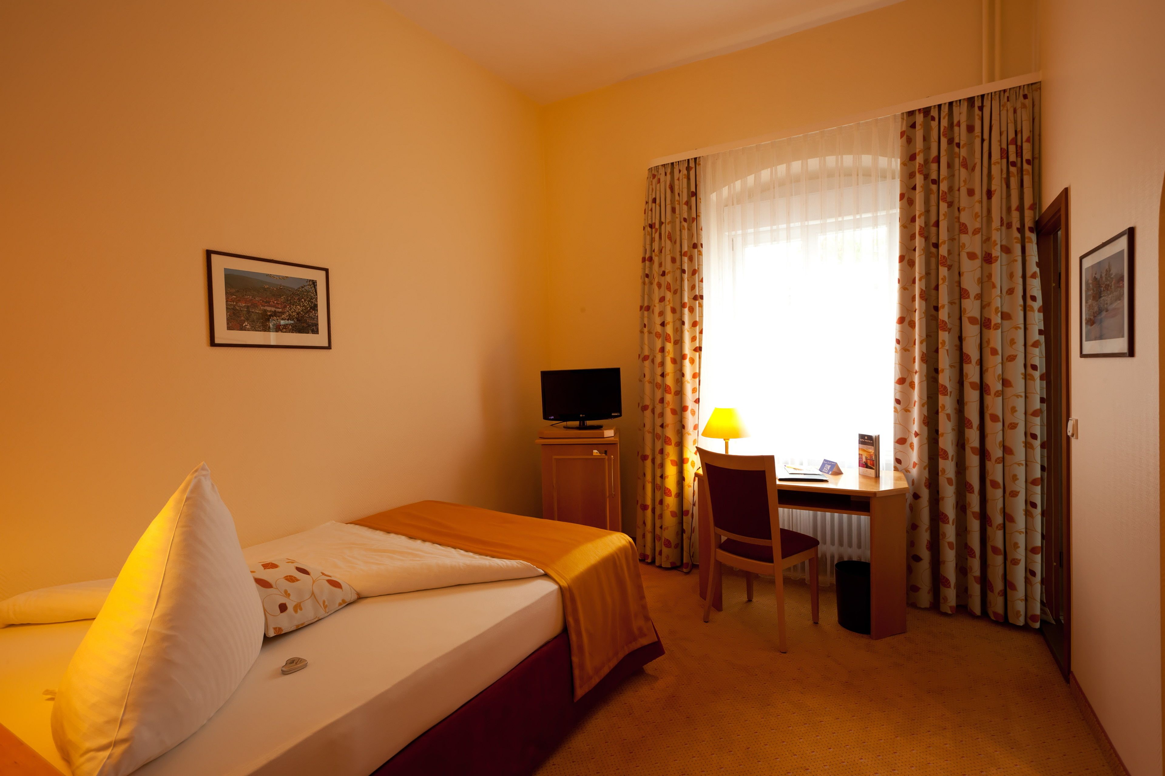 A hotel room with a bed, a desk with a chair, a television, and a window with curtains.