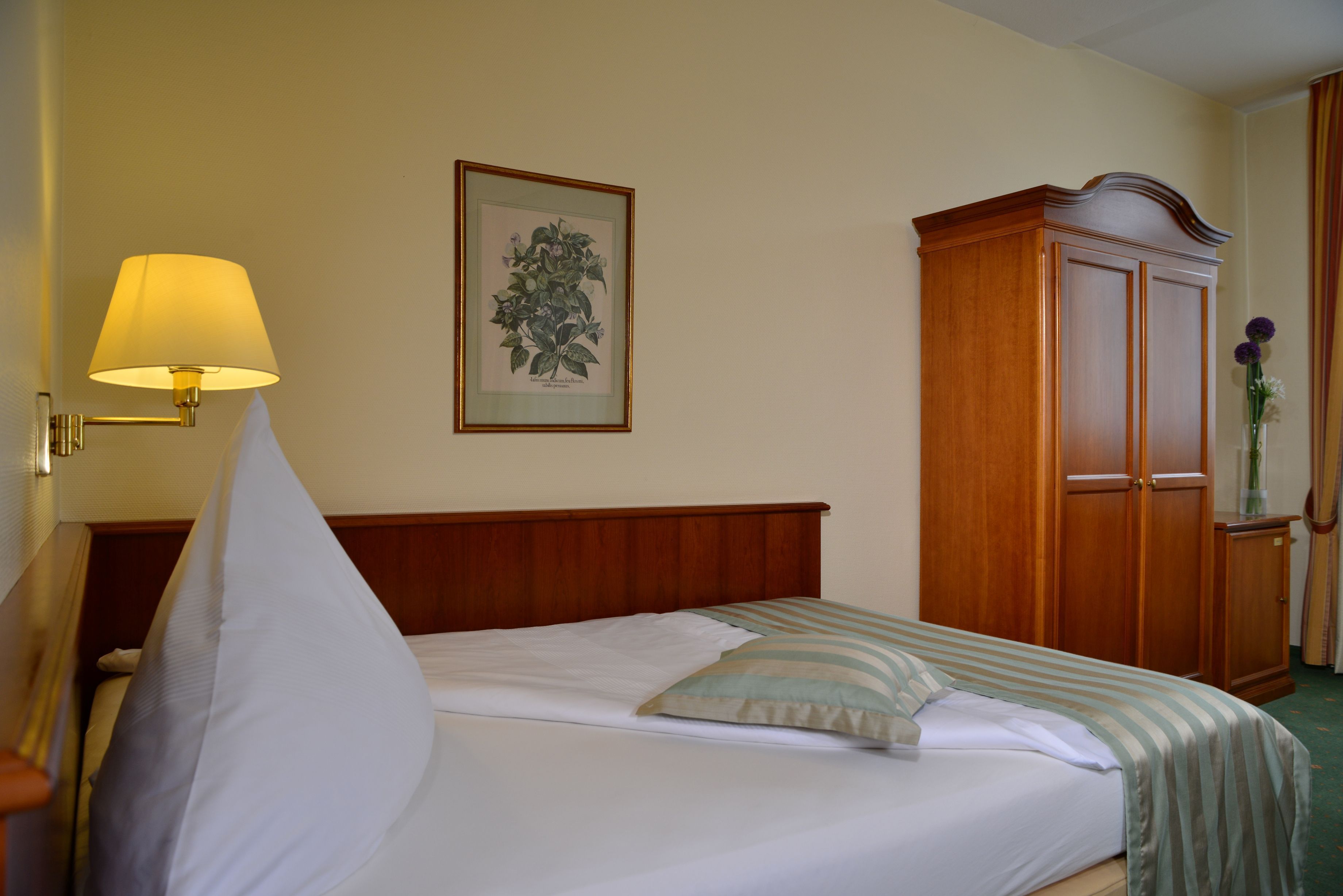 A neatly made bed with a pillow, a bedside lamp, a framed botanical illustration, and a wooden wardrobe.