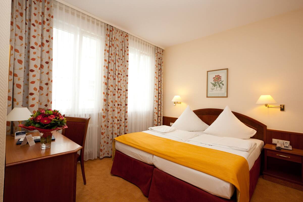 A hotel room with a bed, a desk with a lamp, a chair, and a nightstand with a phone.