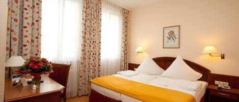 A hotel room with a bed, a desk with a lamp, a chair, and a nightstand with a phone.