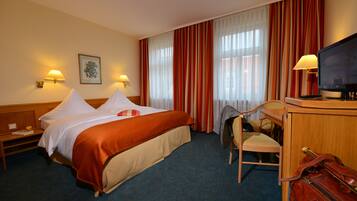 Ein Hotelzimmer mit einem Bett, zwei Nachttischlampen, einem Fernseher, einem Schreibtisch mit Stuhl und einem Koffer.