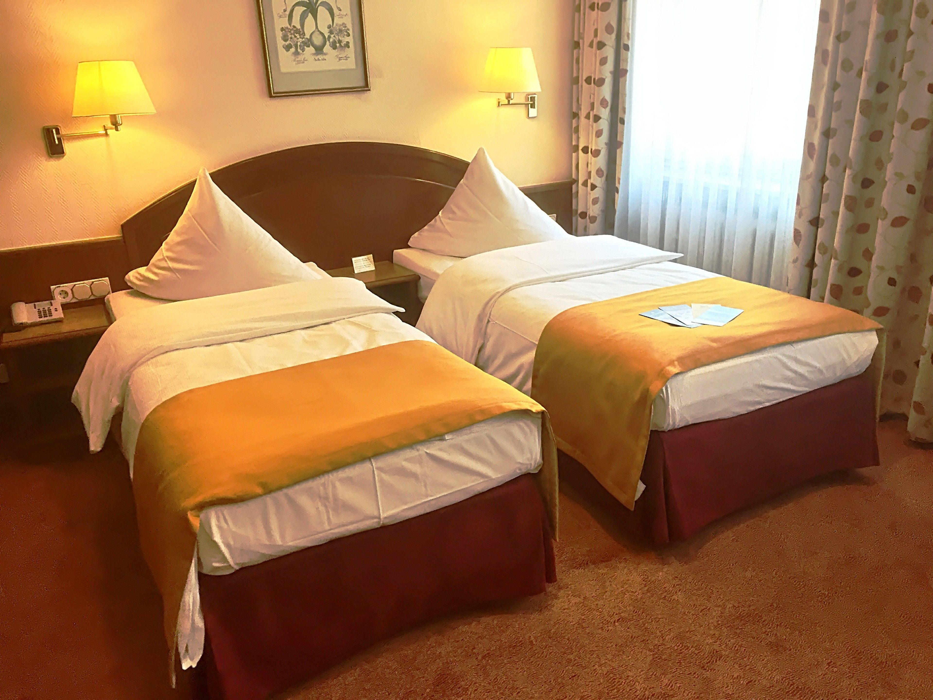 A hotel room with two beds, each with a headboard, white linens, and a red skirted bed base. There are two bedside tables with lamps, a framed picture on the wall, and a window with curtains.