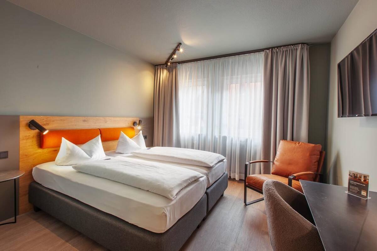 A hotel room with a double bed, a desk with a chair, a TV on the wall, and a window with curtains.
