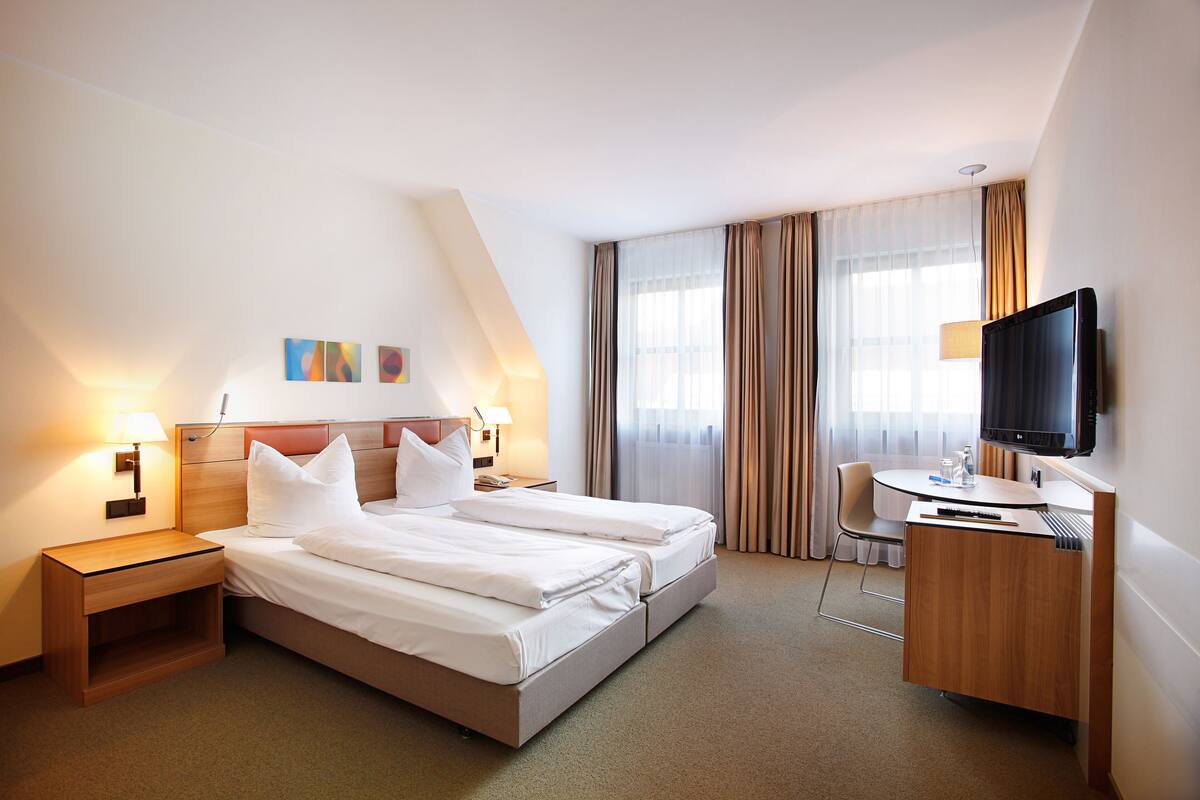 A hotel room with a bed, bedside tables, a desk, a chair, a TV, and curtains.