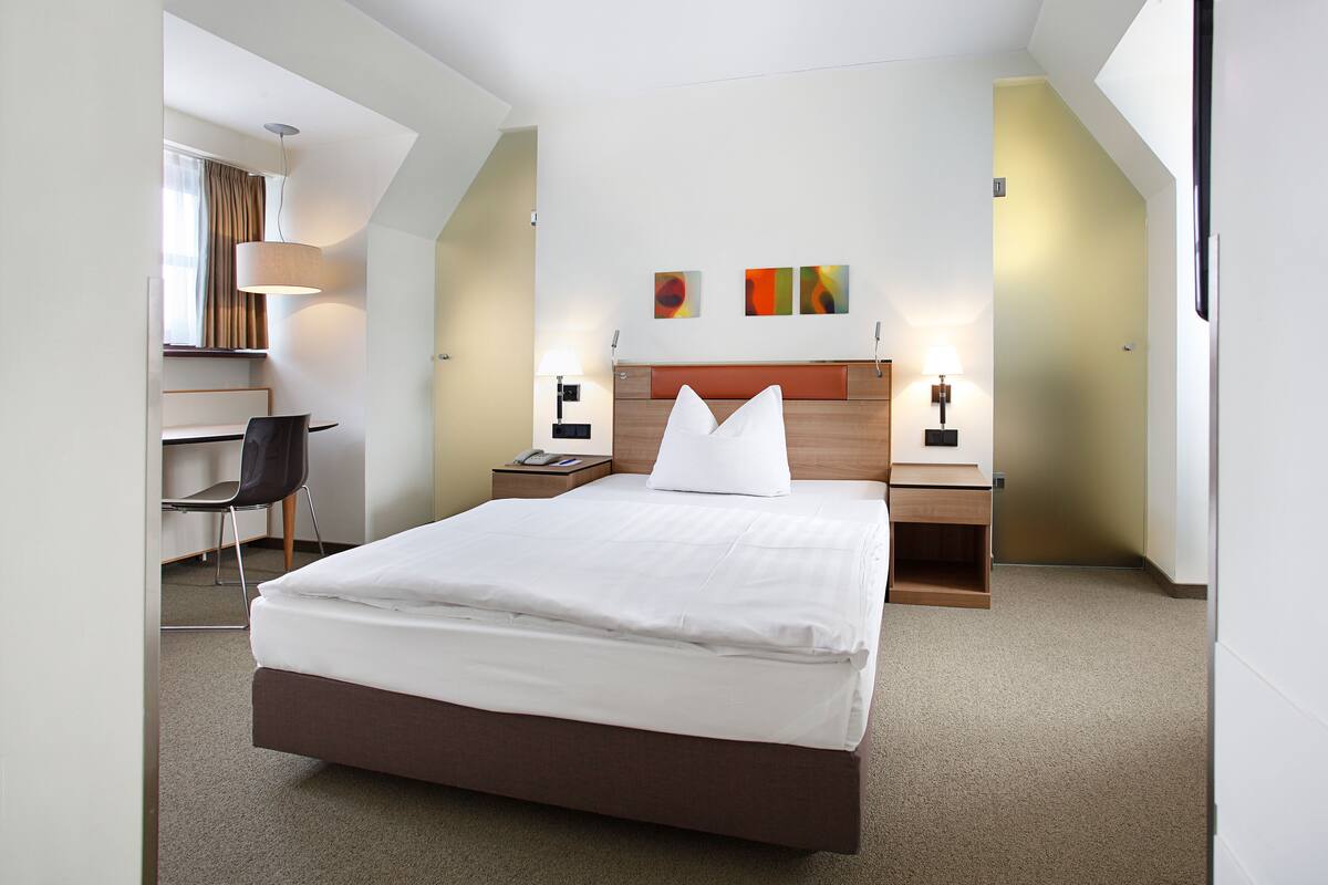A hotel room with a large bed, a desk, a chair, and two bedside tables with lamps.