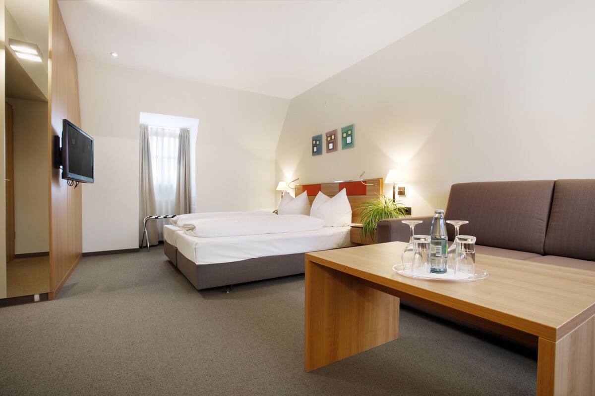 A hotel room with a bed, a sofa, a TV mounted on the wall, and a table with a tray of glasses and a bottle.