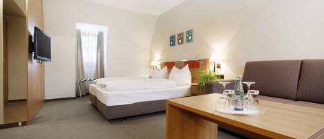 Una habitación de hotel con una cama, un sofá, un televisor montado en la pared y una mesa con una bandeja de vasos y una botella.