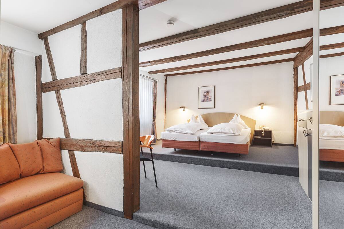 A room with exposed wooden beams, a sofa, a bed with white bedding, a chair, and a nightstand with a lamp.