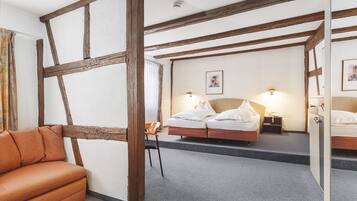 Una habitación con vigas de madera a la vista, un sofá, una cama con ropa blanca, una silla y una mesita de noche con lámpara.