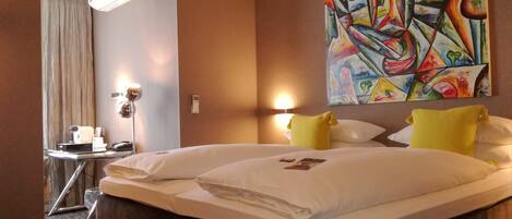 Habitación de hotel con una cama grande, dos almohadas, una mesita de noche con lámpara, un cuadro en la pared y una ventana con cortinas.