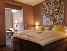 Habitación de hotel con una cama grande, dos almohadas, una mesita de noche con lámpara, un cuadro en la pared y una ventana con cortinas.