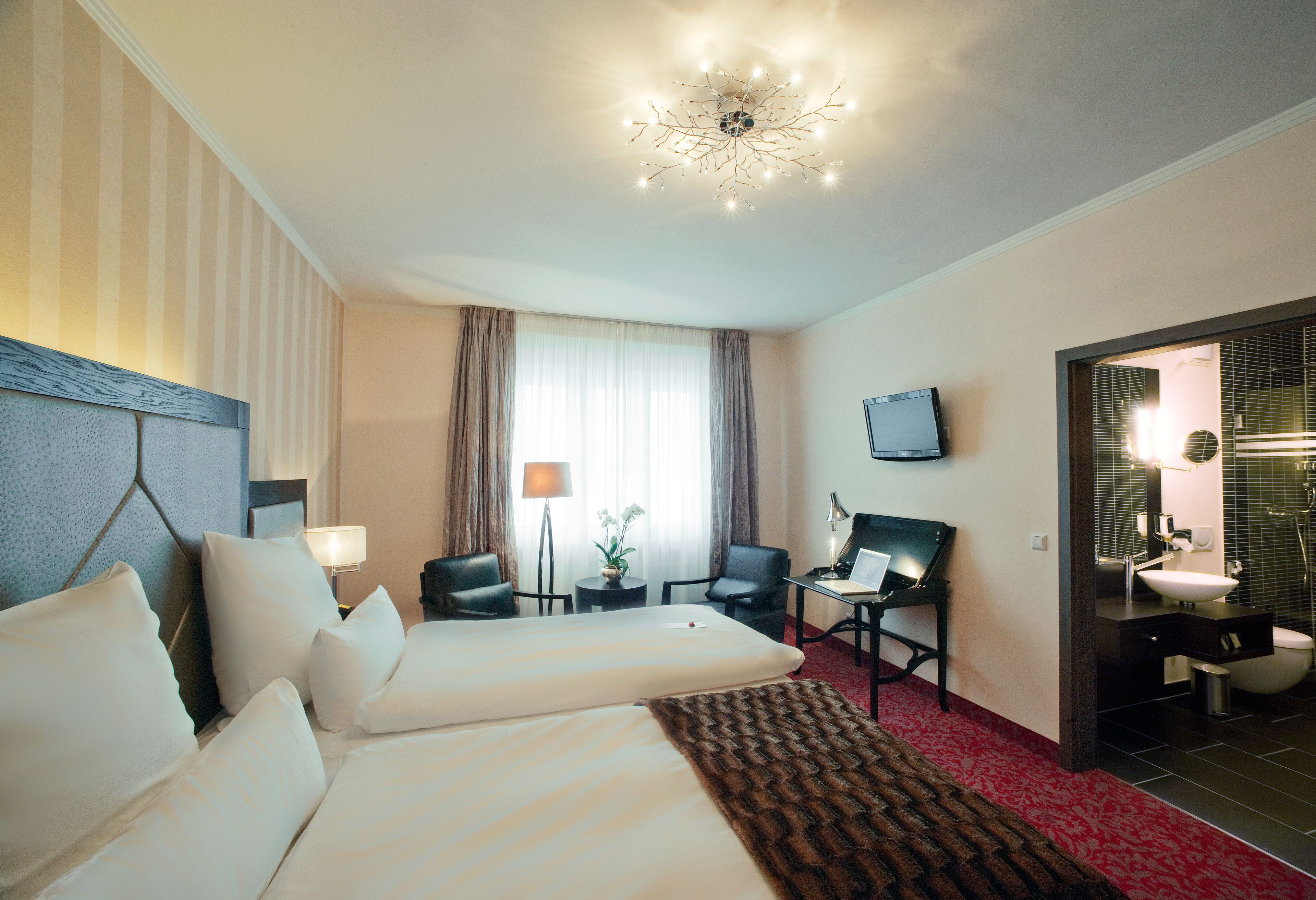 Quarto de hotel com duas camas, uma escrivaninha com cadeira, televisão e banheiro visível através de uma porta aberta.