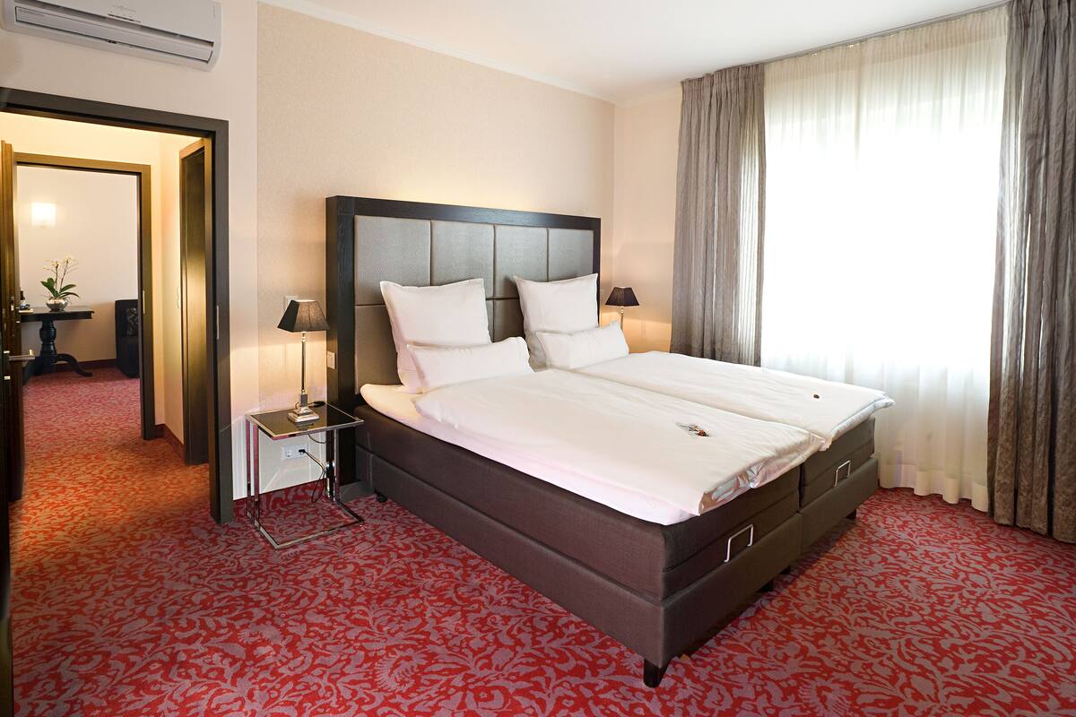 A hotel room with a large bed, a nightstand, a lamp, a mirror, and a view of the outdoors through a window with curtains.