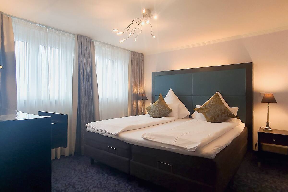 A hotel room with a bed, bedside tables, a desk, and a chandelier.