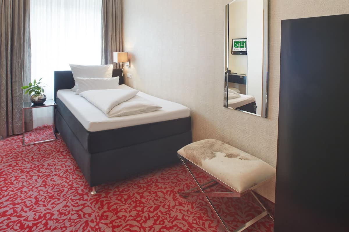 A hotel room with a bed, a bedside table with a lamp, a small stool, a mirror, and a ceiling light fixture.