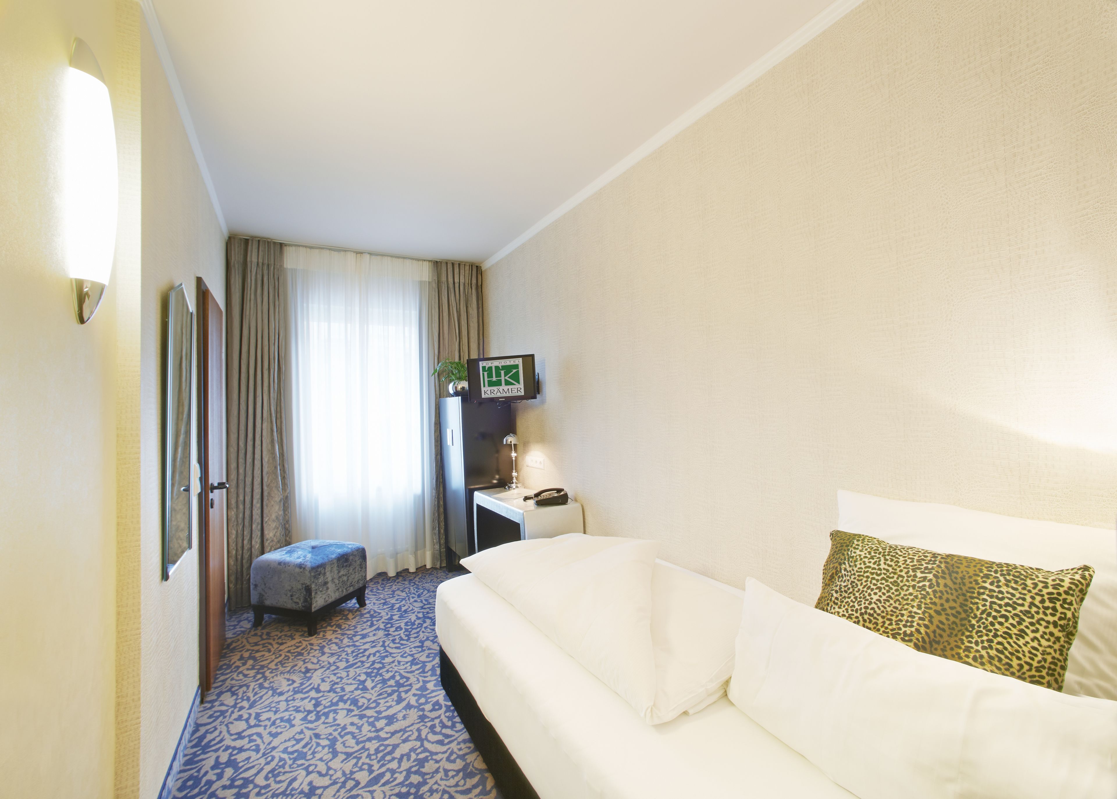 A hotel room with a white sofa, a leopard print pillow, a small table with a lamp, a TV on a stand, and a patterned carpet.