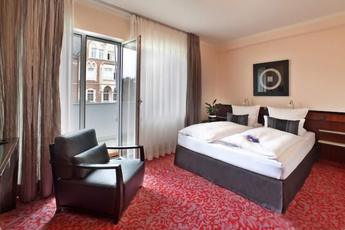 A hotel room with a large bed, a chair, a desk, and a view of a building through the window.