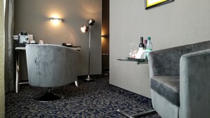 Habitación de hotel con una butaca de mármol, un escritorio con cafetera, una lámpara de pie, un cuadro en la pared y un pequeño estante con botellas.