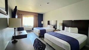 A hotel room with two beds, a desk, and a chair. There is a window with curtains, a lamp, and a phone on the nightstand.