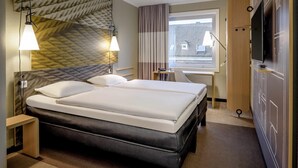 A hotel room with a bed, bedside lamps, a desk, a television, and a window with a view of buildings. - ibis Muenster City (Münster)
