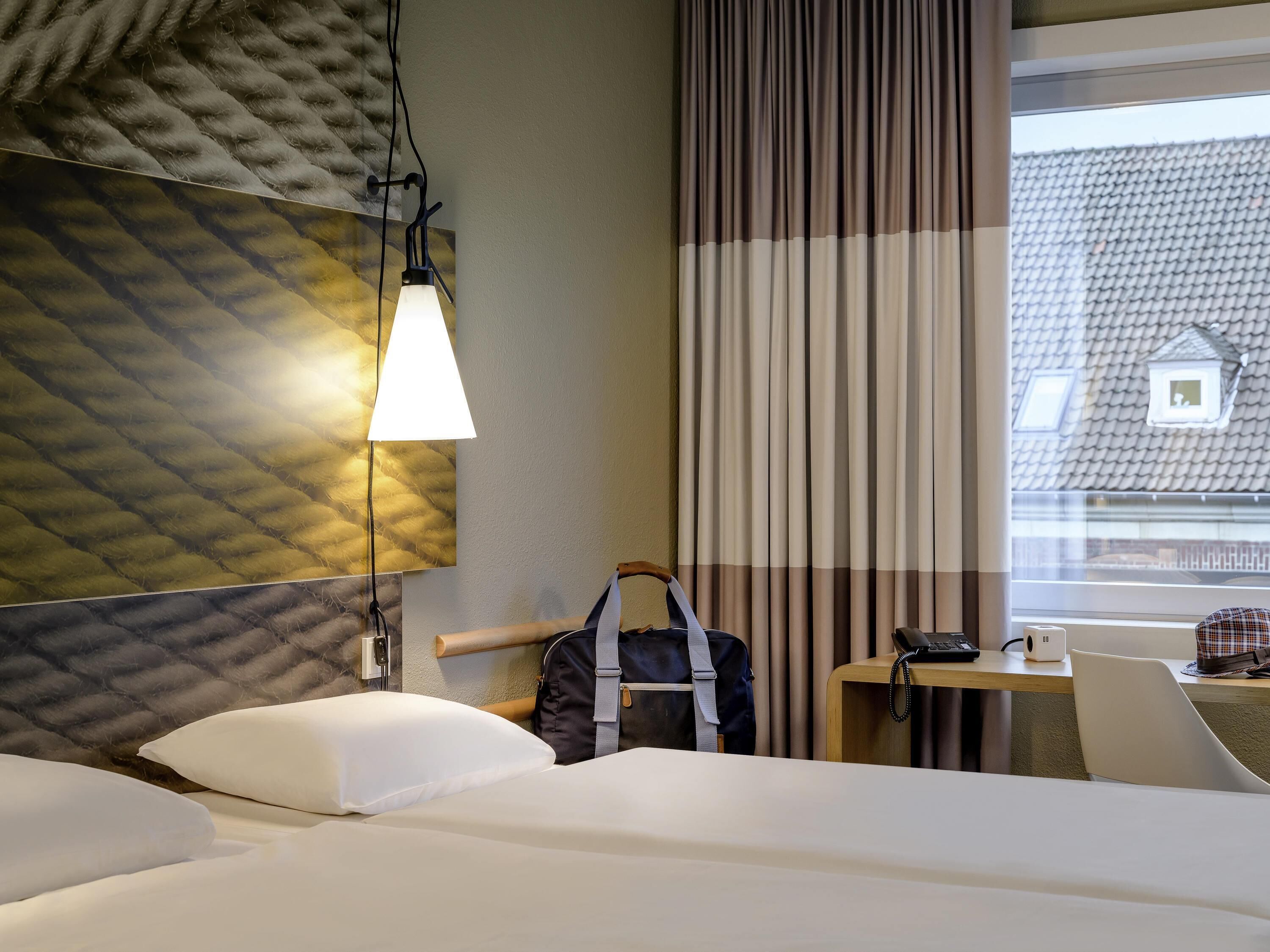 A hotel room with a bed, a desk with a telephone, a hanging bag, and a window with curtains.