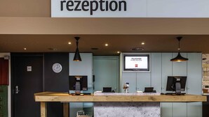 A reception desk with a clock, a digital screen displaying a welcome message, and a green bag on the floor. - ibis Muenster City (Münster)