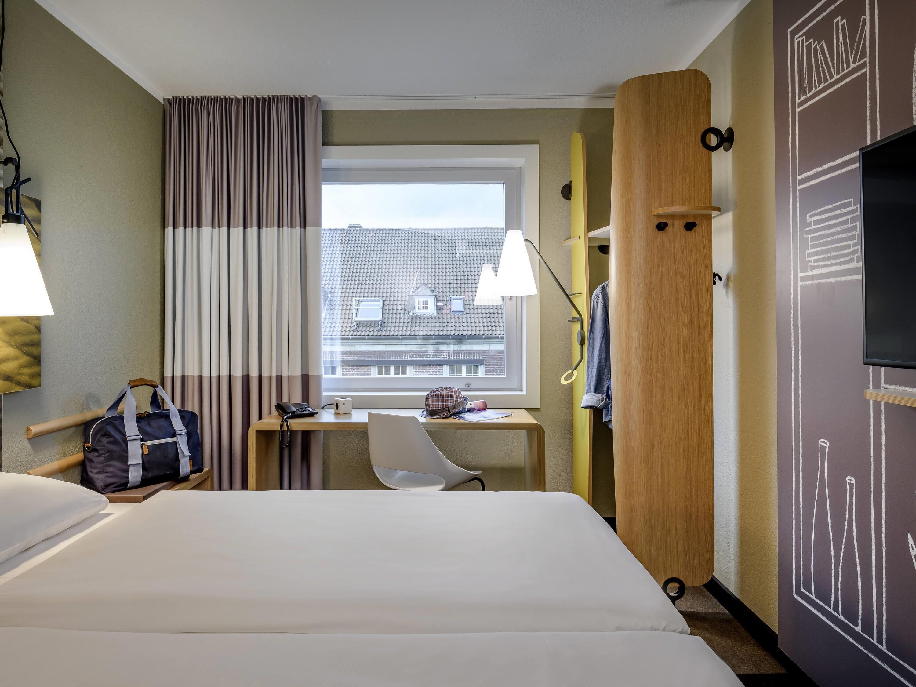 A hotel room with a bed, desk, chair, and a view of buildings through the window.