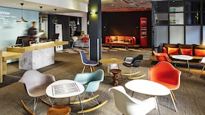 A modern lobby with a reception desk, seating area, and a red information stand. - ibis London Stratford (London)