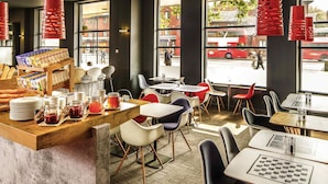 A modern cafe interior with a counter displaying pastries, a coffee machine, and a variety of drinks. There are several tables and chairs arranged for seating. - ibis London Stratford (London)