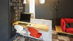 A modern office space with a wooden desk, a laptop, a white chair, and a red chair. There is a red sofa in the background. - ibis London Stratford (London)