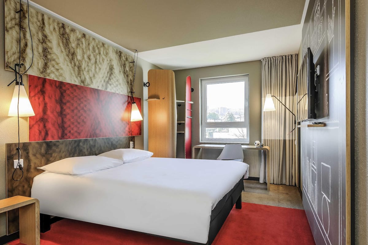 A hotel room with a bed, bedside lamps, a TV, a window, and a red carpet.