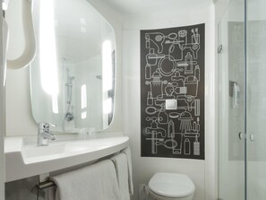 A bathroom with a white sink, mirror, and toilet, and a shower with a glass door. - ibis London Stratford (London)
