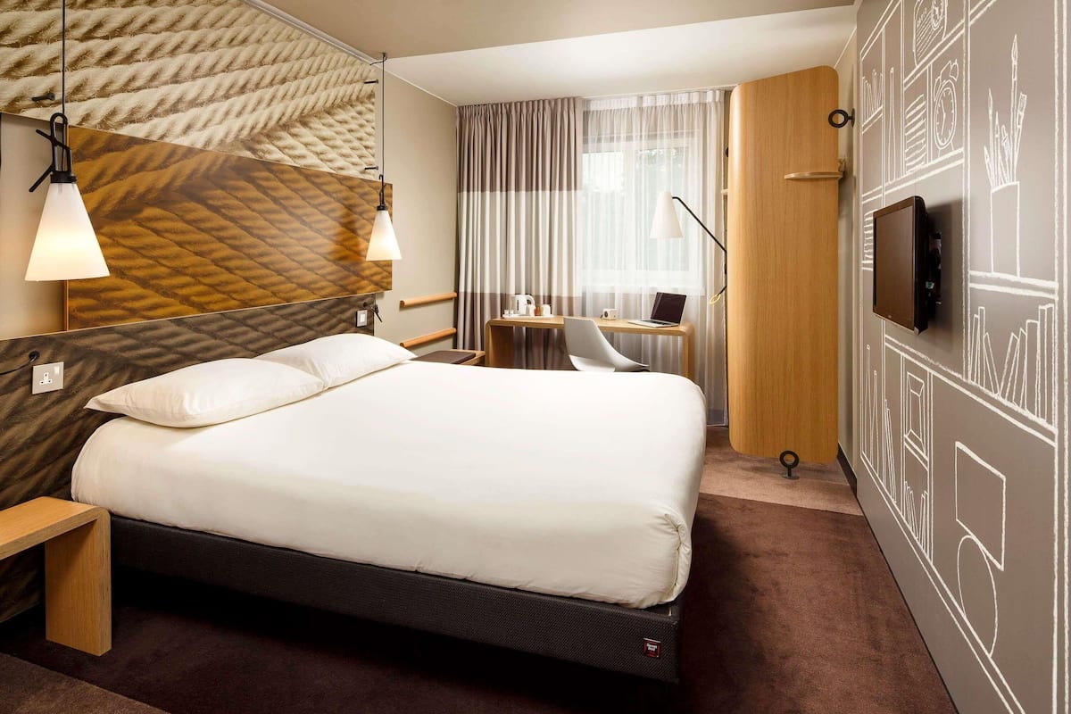 A modern hotel room with a large bed, a desk with a laptop, a TV, and a unique textured wall design.
