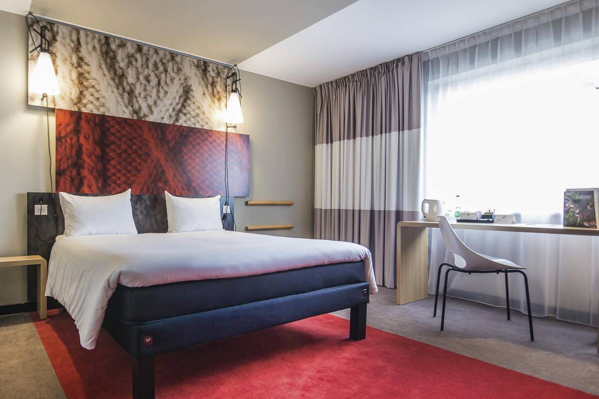 A hotel room with a bed, desk, chair, and large windows with curtains.