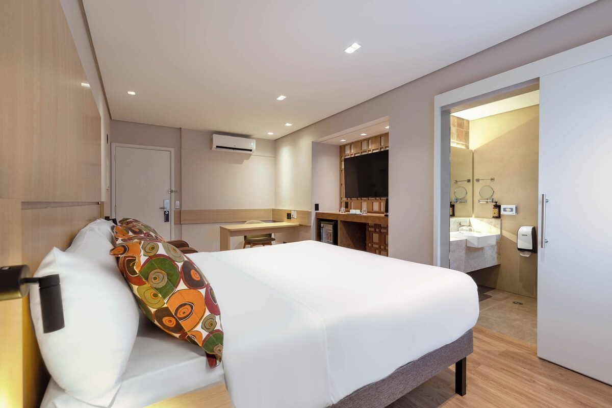 Habitación de hotel con cama, escritorio, espejo y baño visible a través de una puerta abierta.