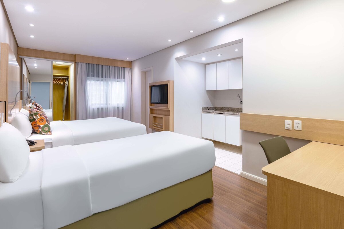 A hotel room with two beds, a desk, a TV, a kitchenette, and a walk-in closet.