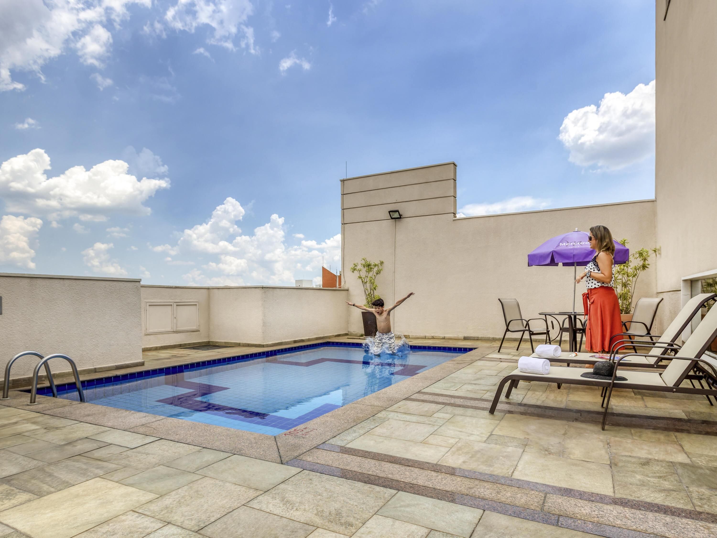 A rooftop pool with a clear blue sky, a lounge area with chairs and an umbrella, and a building with a flat roof.
