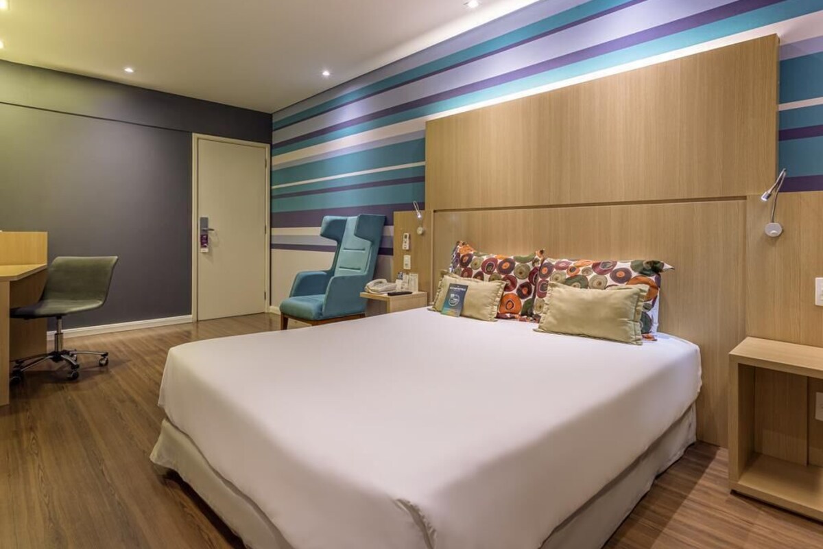 A hotel room with a large bed, a desk, a chair, and a wall with horizontal stripes.