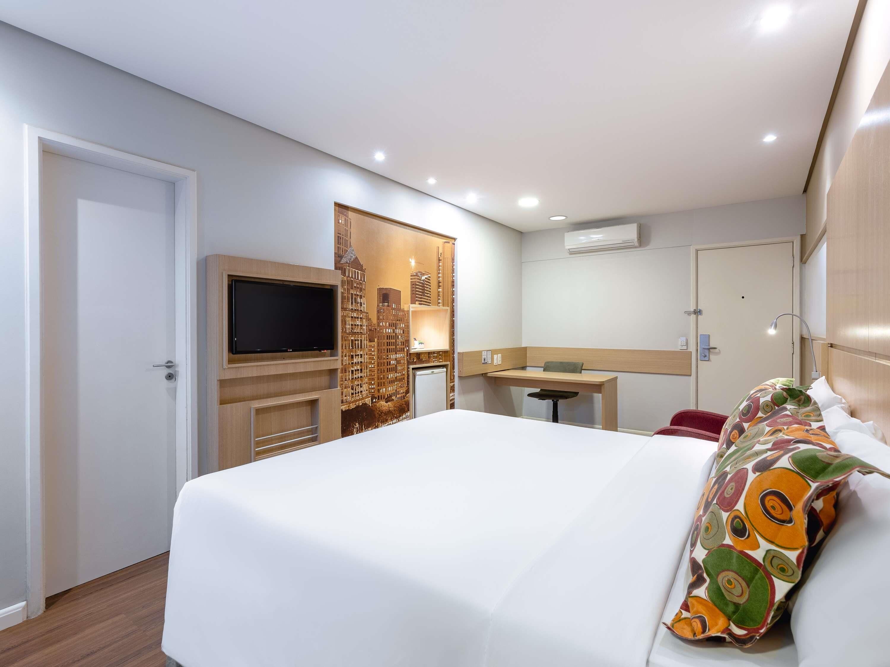Quarto de hotel com cama, escrivaninha, televisão e ar condicionado.