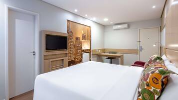 Quarto de hotel com cama, escrivaninha, televisão e ar condicionado.