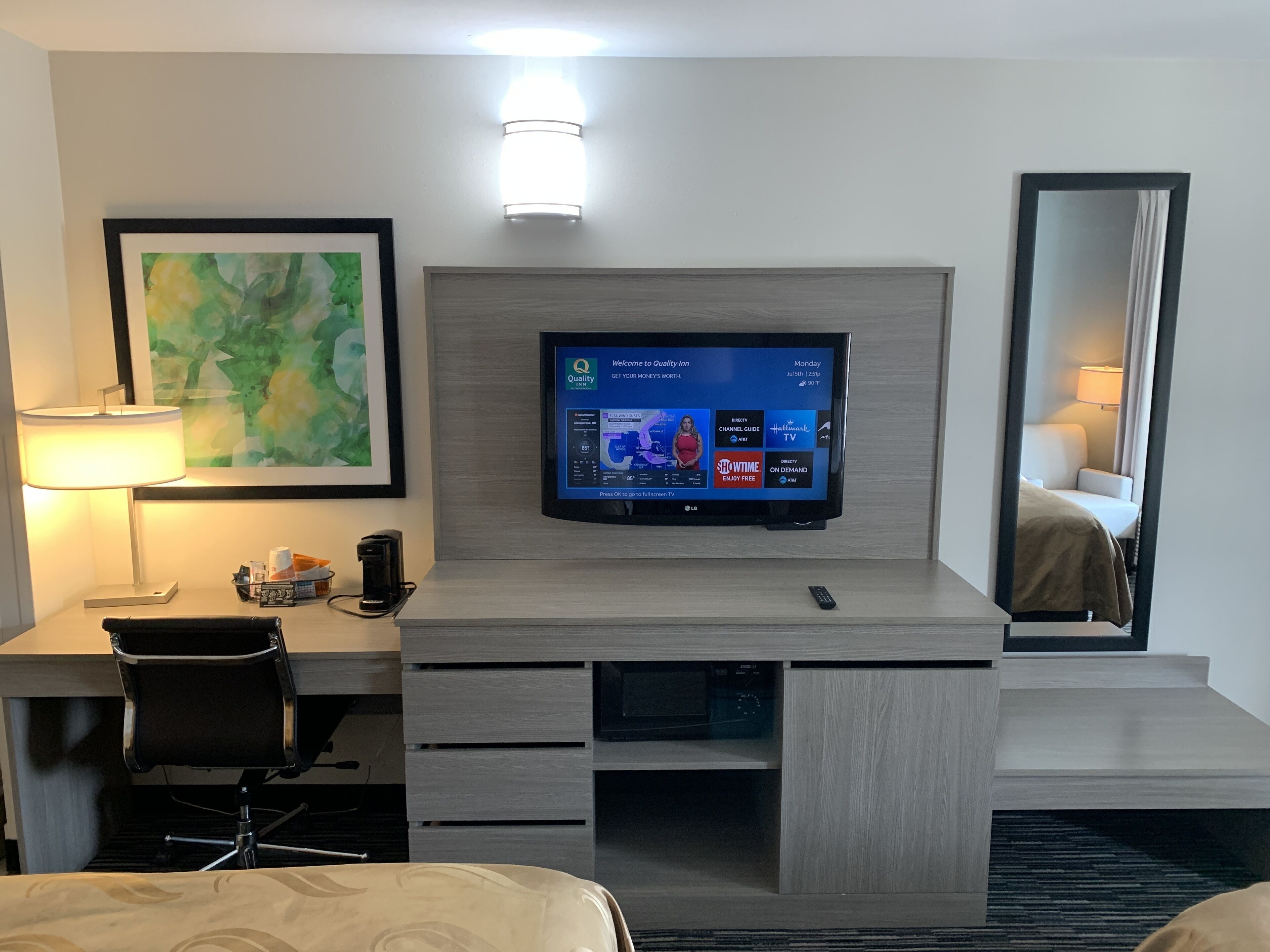 Une chambre d’hôtel équipée d’un téléviseur à écran plat fixé au mur, d’un bureau avec une lampe, d’une chaise et d’un lit avec une tête de lit.