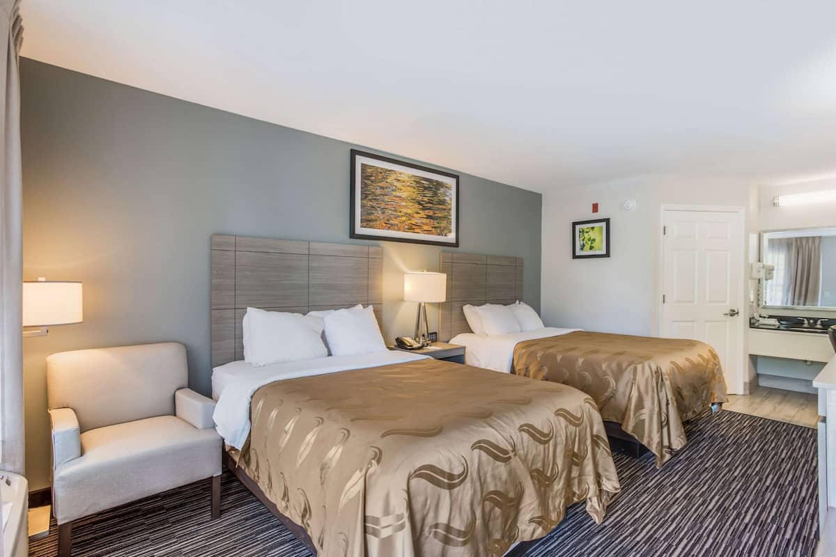 A hotel room with two beds, a desk, a chair, a lamp, and a framed picture on the wall.