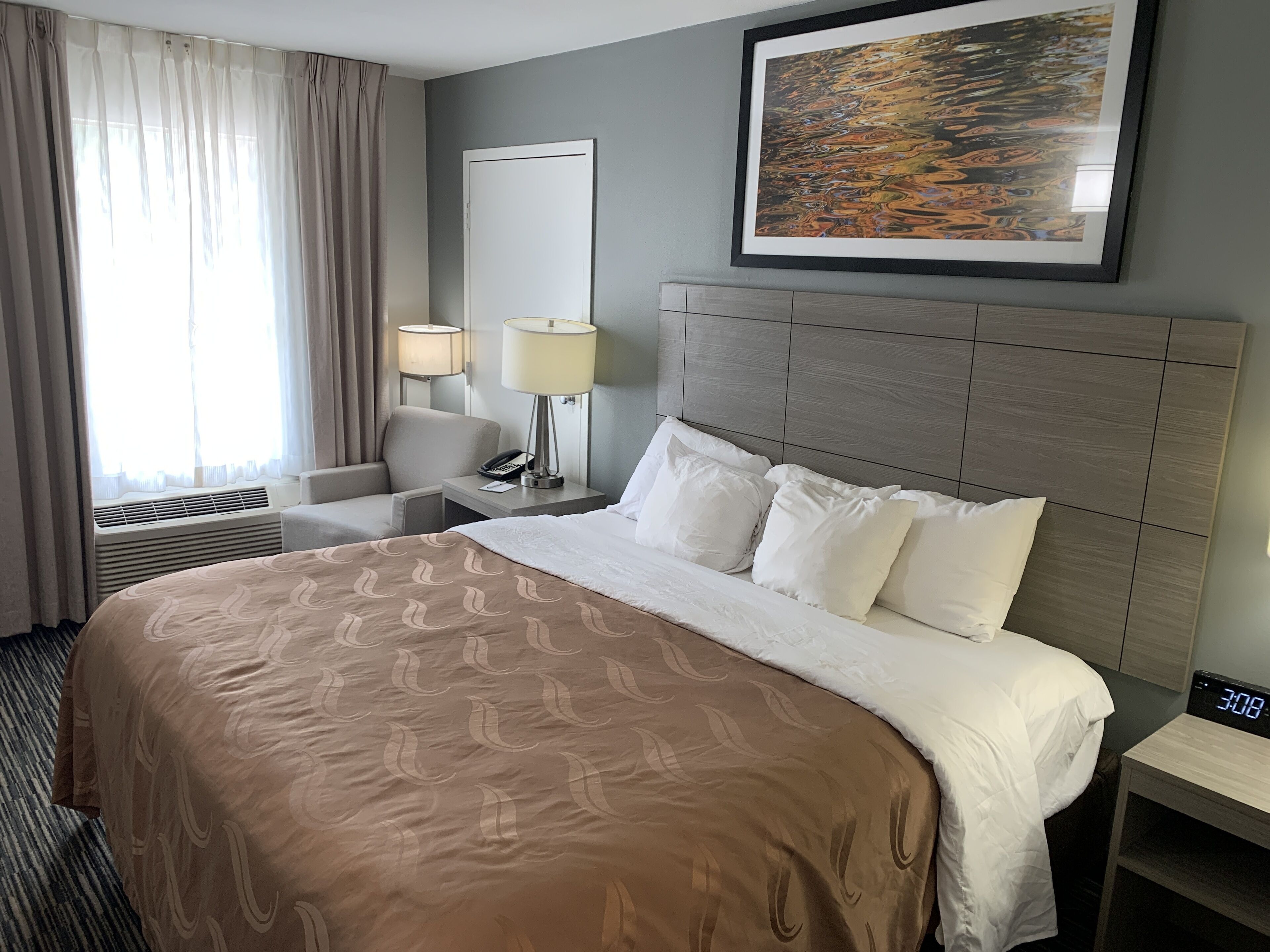 A hotel room with a large bed, a headboard, a bedside table with a lamp, a chair, a window with curtains, and a framed picture on the wall.