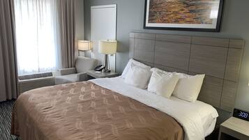 Una habitación de hotel con una cama grande, un cabecero, una mesita de noche con lámpara, una silla, una ventana con cortinas y un cuadro en la pared.