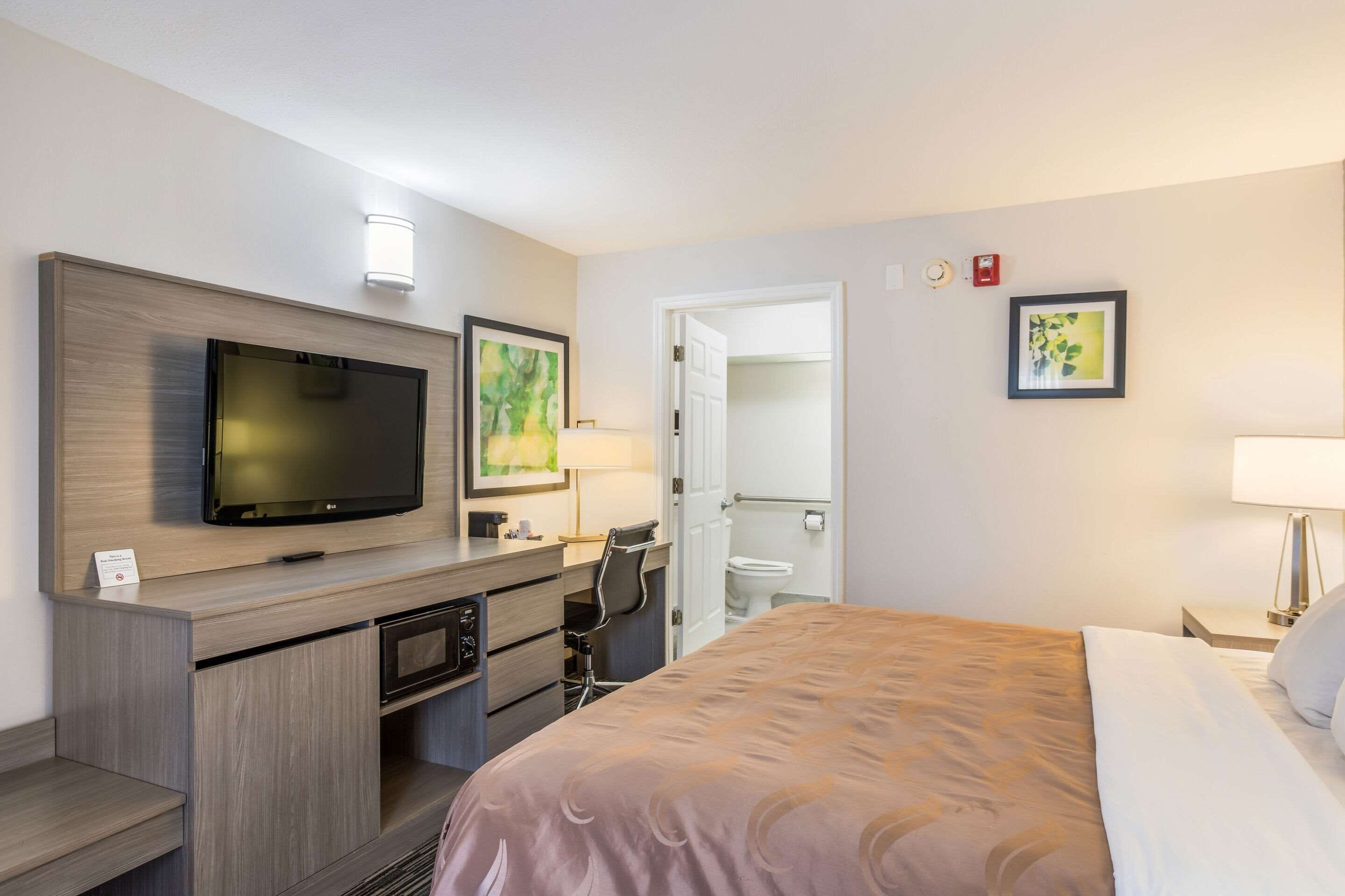 A hotel room with a bed, a desk with a TV, a microwave, and a chair. There is a bathroom visible through an open door.