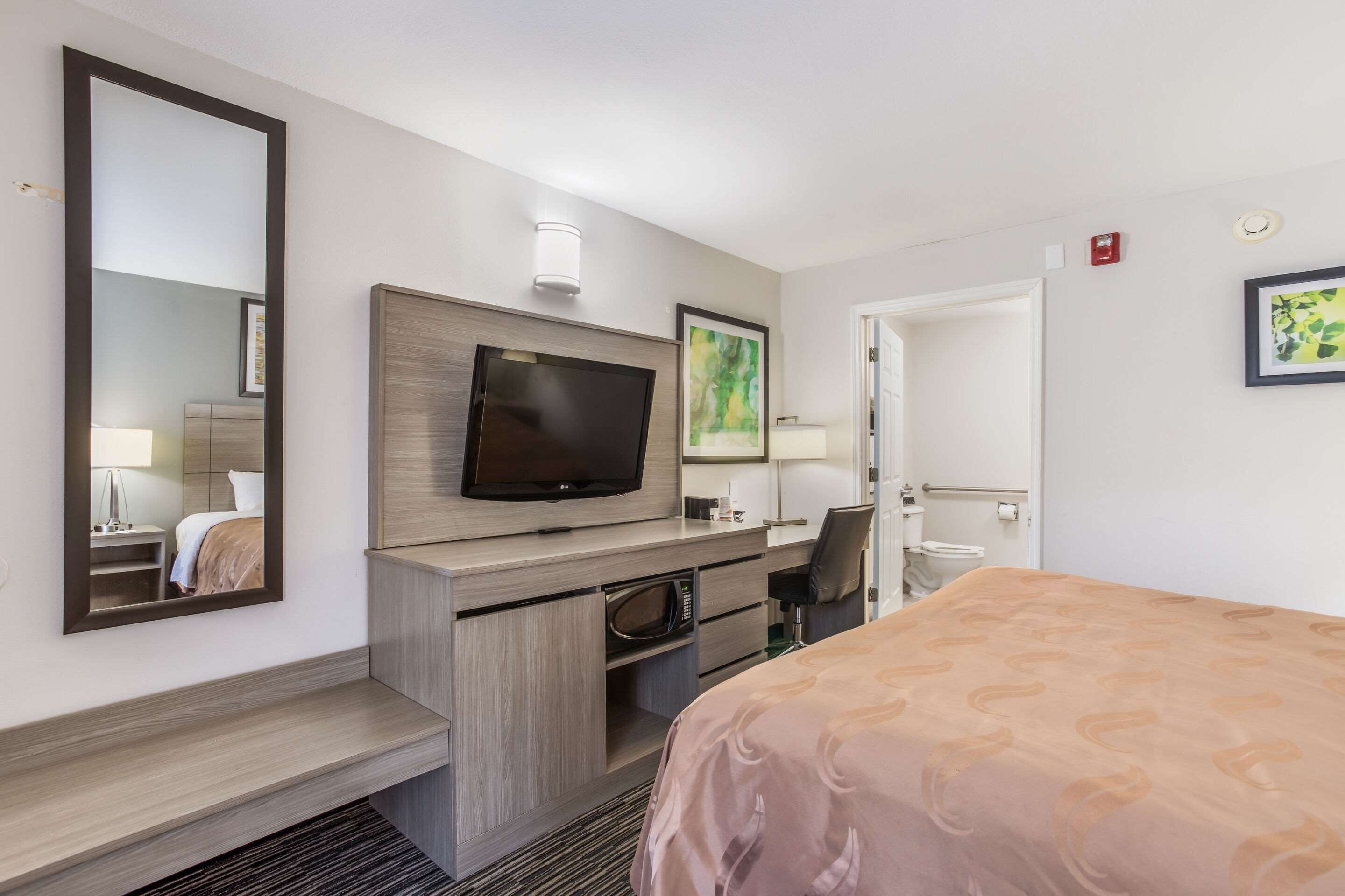 A hotel room with a bed, a TV, a desk with a chair, a microwave, a mirror, and a door leading to a bathroom.