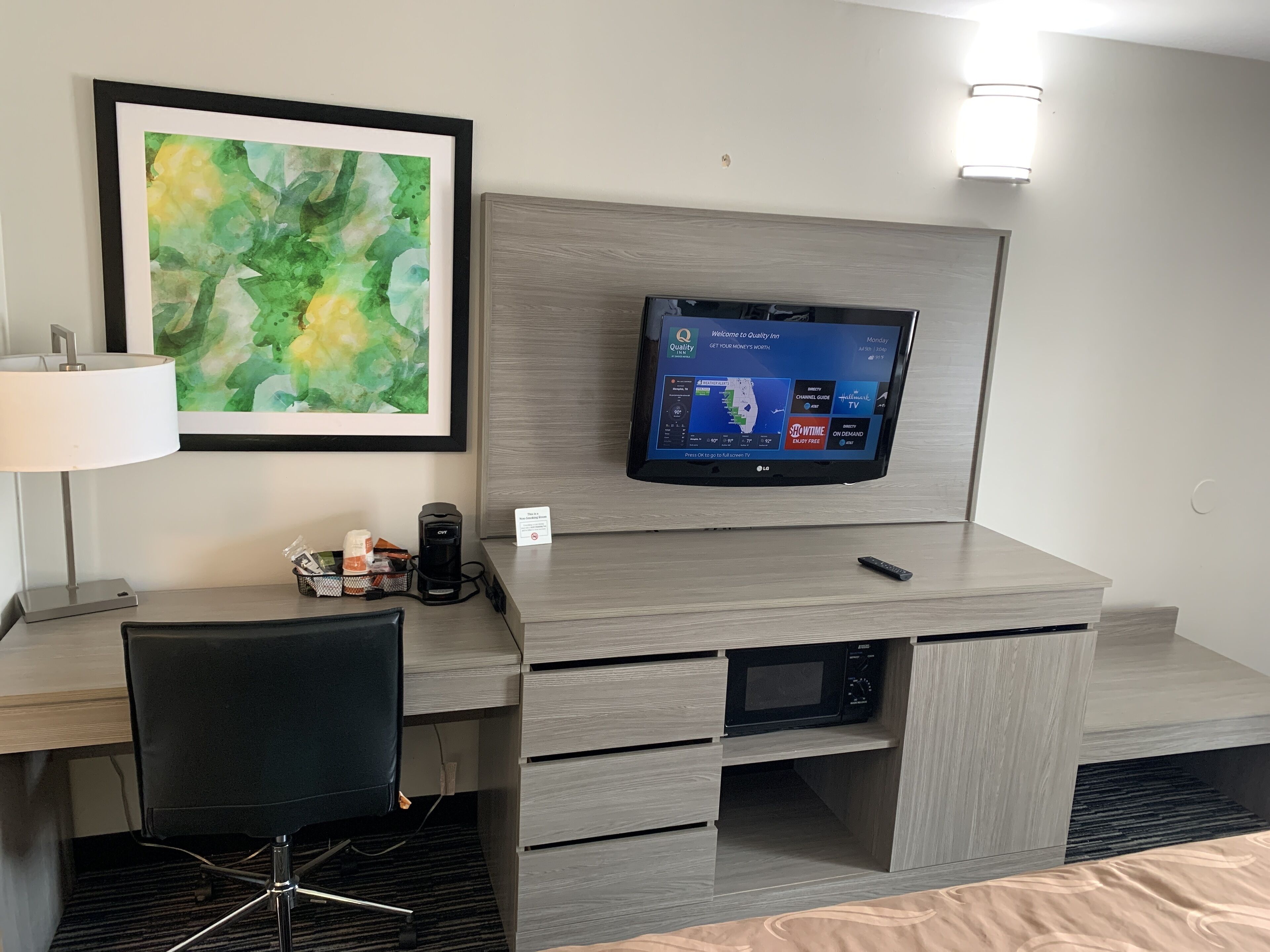 A hotel room with a flat-screen TV mounted on a wooden wall, a desk with a chair, a lamp, and a coffee maker.