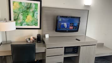 A hotel room with a flat-screen TV mounted on a wooden wall, a desk with a chair, a lamp, and a coffee maker.
