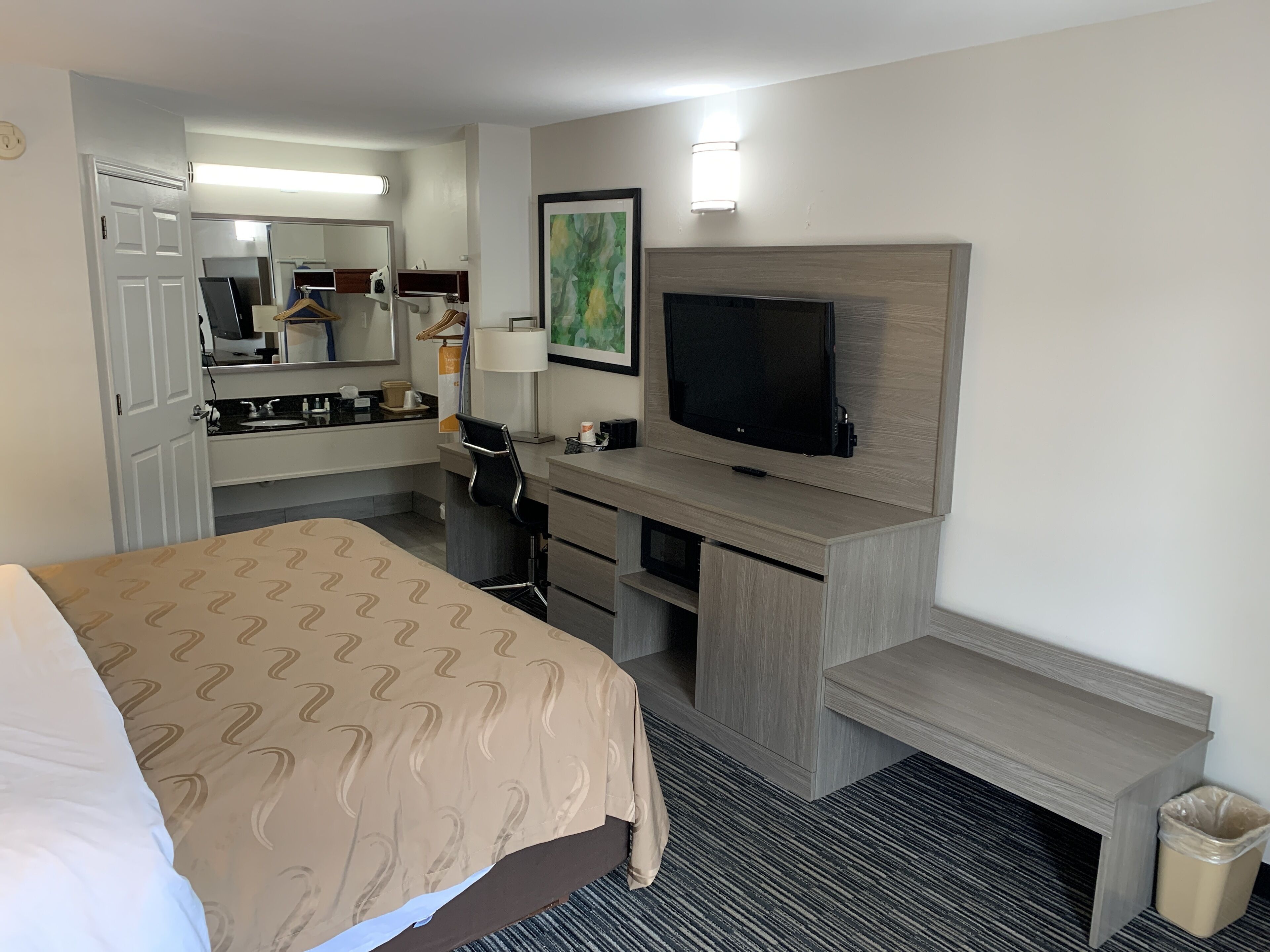 A hotel room with a bed, a desk with a television, a mirror, a trash bin, and a painting on the wall.